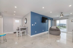 A modern living room with a blue accent wall, a flat-screen TV, a ceiling fan, and a large bean bag chair. - Lakefront Gem | Cozy Stay w/ Dock + Sunset Views (Sodus Point)