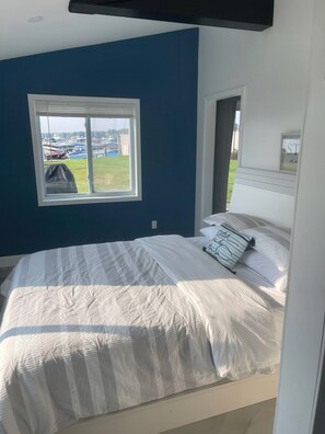 A bedroom with a bed, a window with a view of boats, and a white headboard. - Lakefront Gem | Cozy Stay w/ Dock + Sunset Views (Sodus Point)