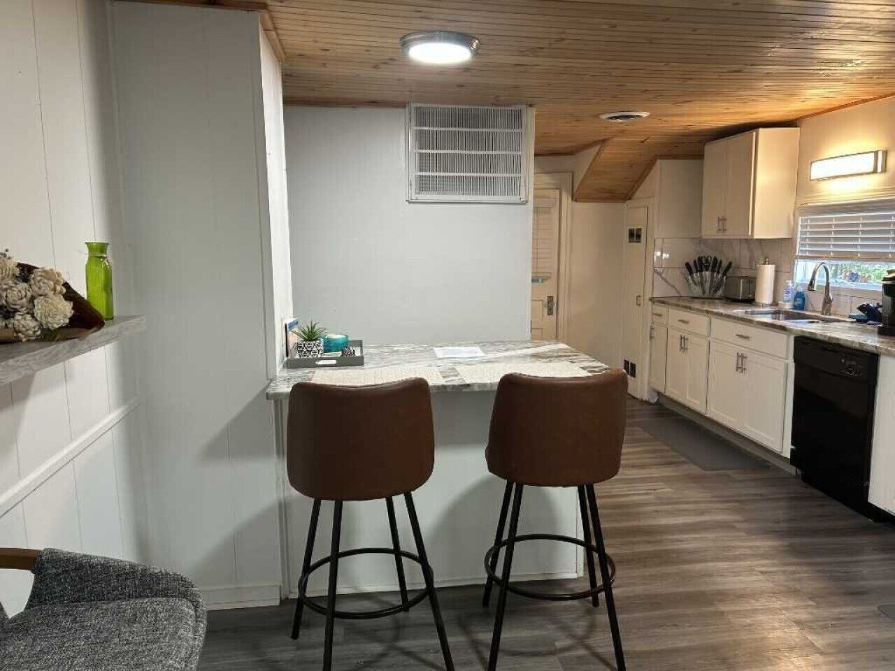 Une cuisine avec un coin bar, un plafond en bois, des meubles blancs et une fenêtre avec des stores.