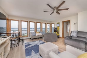 A modern living room with a large window, a ceiling fan, a sofa, a bean bag, and a small table. - Waterfront Sodus Gem w/ Dock, Game Loft, Bay View (Sodus Point)