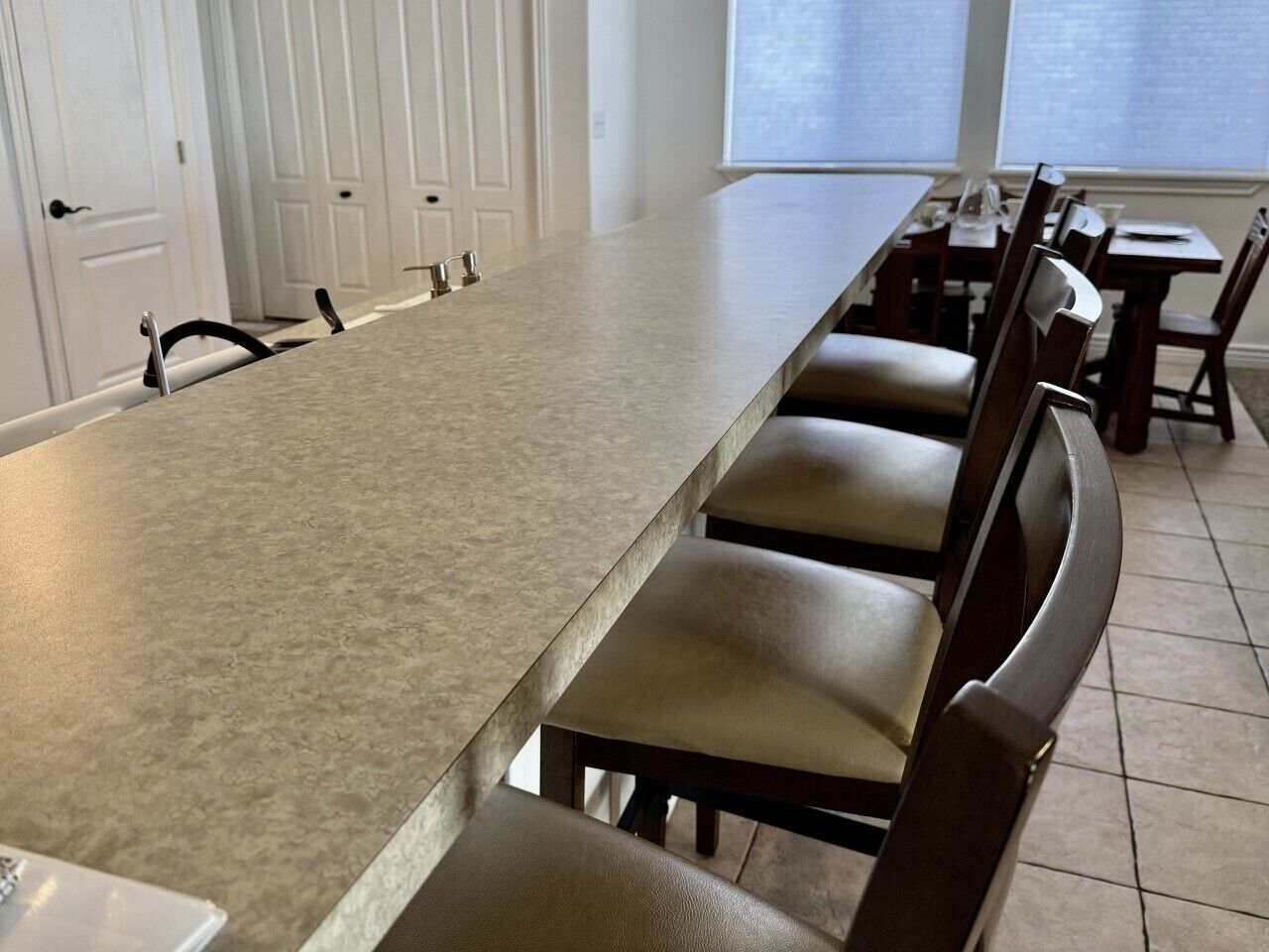 Une longue table à manger entourée de chaises, des portes blanches en arrière-plan et une fenêtre avec des stores.