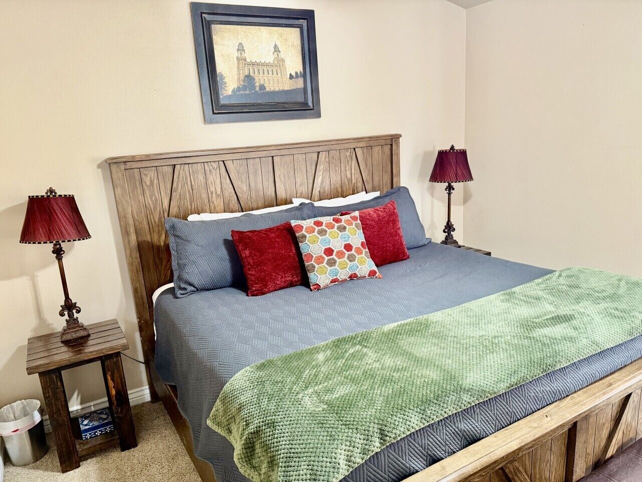 A bedroom with a wooden headboard, a bed with blue bedding and red pillows, a green throw blanket, and two bedside lamps.