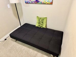 Un sofá cama pequeño de color oscuro con un cojín verde frente a una pared lisa, un enchufe y una obra de arte colorida en la parte superior.