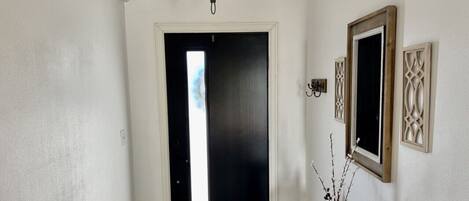 Un pasillo con una puerta negra, una lámpara y un jarrón con ramas sobre una mesa.