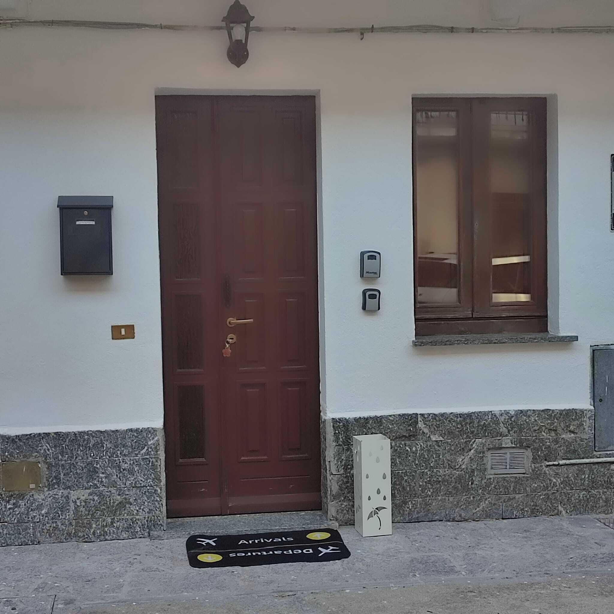 Ein Gebäudeeingang mit brauner Tür, einem Briefkasten, einem Fenster und einer Fußmatte mit An- und Abreisehinweisen.