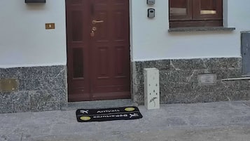 Ein Gebäudeeingang mit brauner Tür, einem Briefkasten, einem Fenster und einer Fußmatte mit An- und Abreisehinweisen.