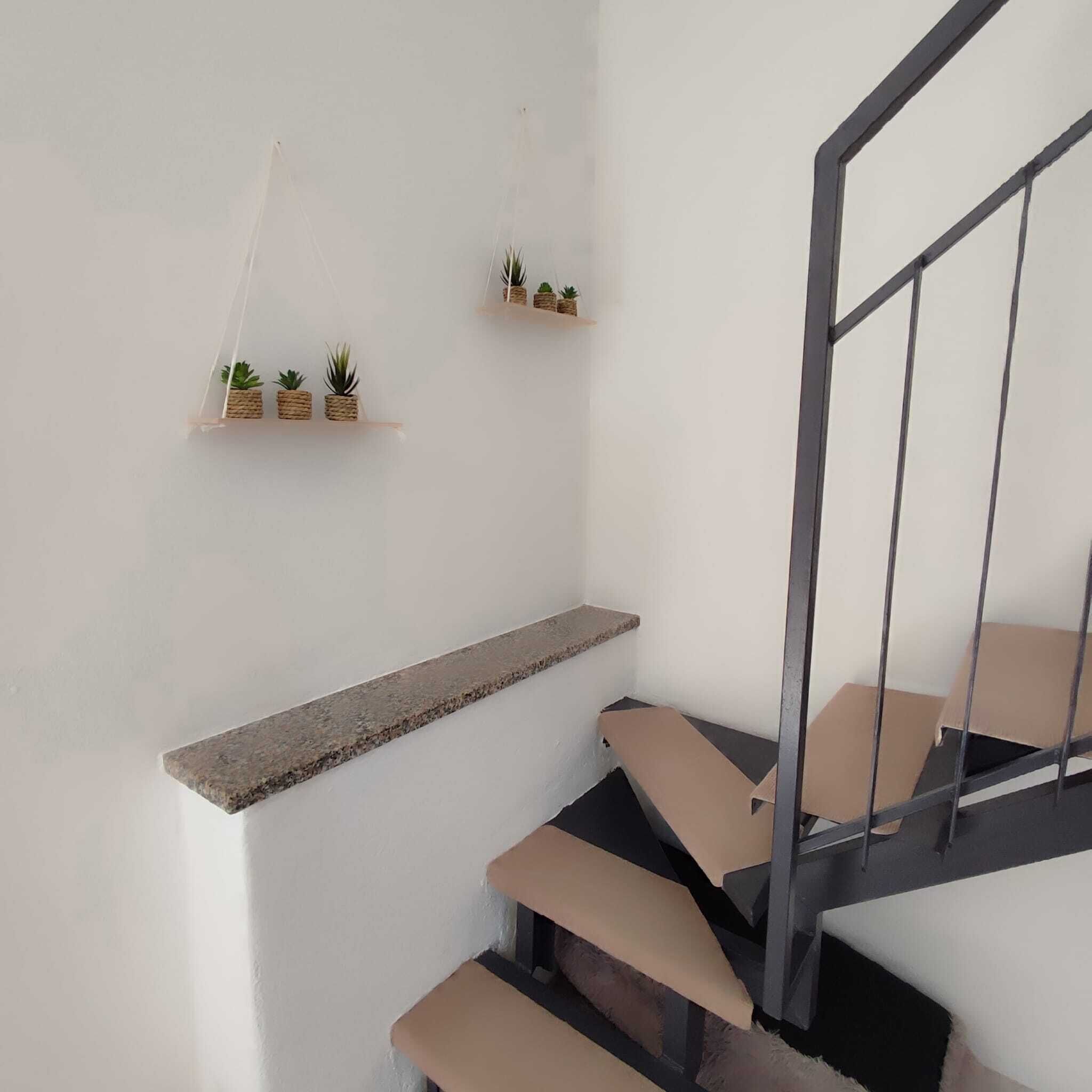 Una scala con ringhiera in legno e corrimano metallico, che conduce a un pianerottolo con uno scaffale su cui sono disposti piccoli vasi di piante.