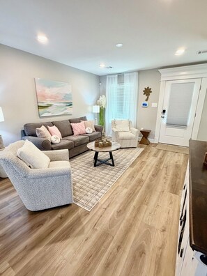 Una sala de estar con sofá, sillón y mesa de centro, un cuadro en la pared y una puerta con ventana.