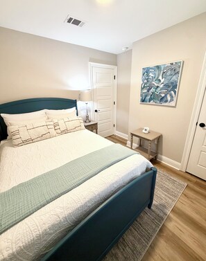 A bedroom with a bed, a nightstand, a lamp, a door, and a painting on the wall. - Gone Coastal •Steps to Beach •Pickleball •Heart of Biloxi (Biloxi)