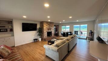 A spacious living room with a fireplace, a large sofa set, a flat-screen TV, and a dining area with a view of the outdoors.