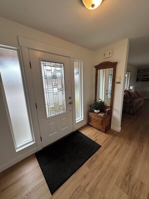 A hallway with a wooden floor, a black doormat, a wooden bench with a plant, and a mirror on the wall.