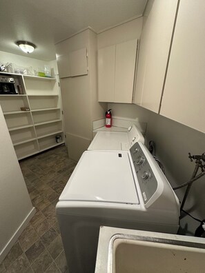 A laundry room with a front-loading washer and dryer, a fire extinguisher, and a shelving unit with various items.