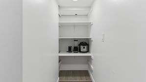 Un couloir étroit aux murs blancs et au plancher en bois. On y trouve trois étagères blanches. Sur l’étagère du haut, il y a une machine à café, un mixeur et une mijoteuse.