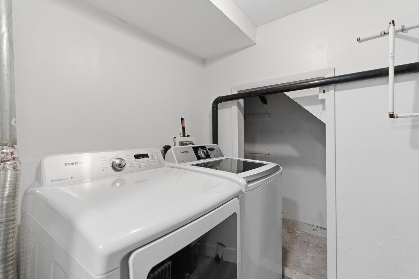 A laundry area with a Samsung washing machine and dryer, a metal pipe, and a white wall.