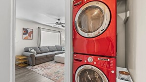 Une buanderie avec un lave-linge et un sèche-linge à chargement frontal rouges, un canapé gris, un ventilateur de plafond et un tapis coloré.