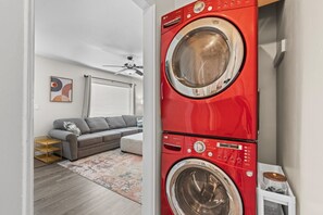 A laundry room with a red front-loading washer and dryer, a gray sofa, a ceiling fan, and a colorful rug. - 1950s Bungalow in Millcreek | Spacious & Central (South Salt Lake)