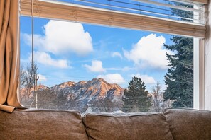 A room with a view of mountains through a window with blinds, a brown sofa, and a curtain. - 1950s Bungalow in Millcreek | Spacious & Central (South Salt Lake)