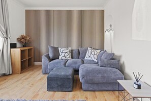A modern living room with a sectional sofa, ottomans, a wooden side table, and a floor lamp. - Modern 3BR Home Perfect for Groups and families (Little Hulton)