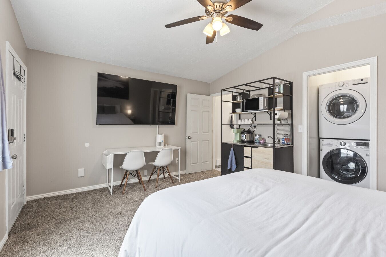 A bedroom with a bed, a desk with chairs, a television, a ceiling fan, and a built-in washer and dryer.