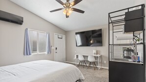 Una camera d'albergo con un letto, una scrivania, una sedia, un televisore, una ventola a soffitto e un frigobar.