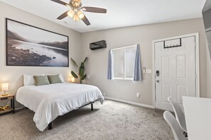 A bedroom with a bed, a ceiling fan, a window with curtains, a door, and a wall-mounted air conditioning unit. - Cozy Millcreek Studio w/ Laundry & Comfort (South Salt Lake)