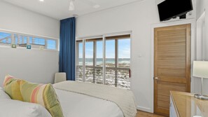 Ein Schlafzimmer mit einem Bett, einem Fenster mit Blick auf den Strand, einer Holztür und einem an der Wand befestigten Fernseher.
