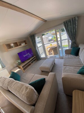 A living room with a sofa set, a television, a wooden shelf, and a sliding glass door leading to an outdoor balcony. - Luxury caravan on a 5 star holiday park (Llanrug)
