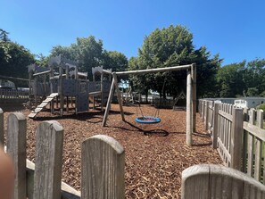 A playground with a wooden swing set and a slide, surrounded by a wooden fence. - Luxury caravan on a 5 star holiday park (Llanrug)