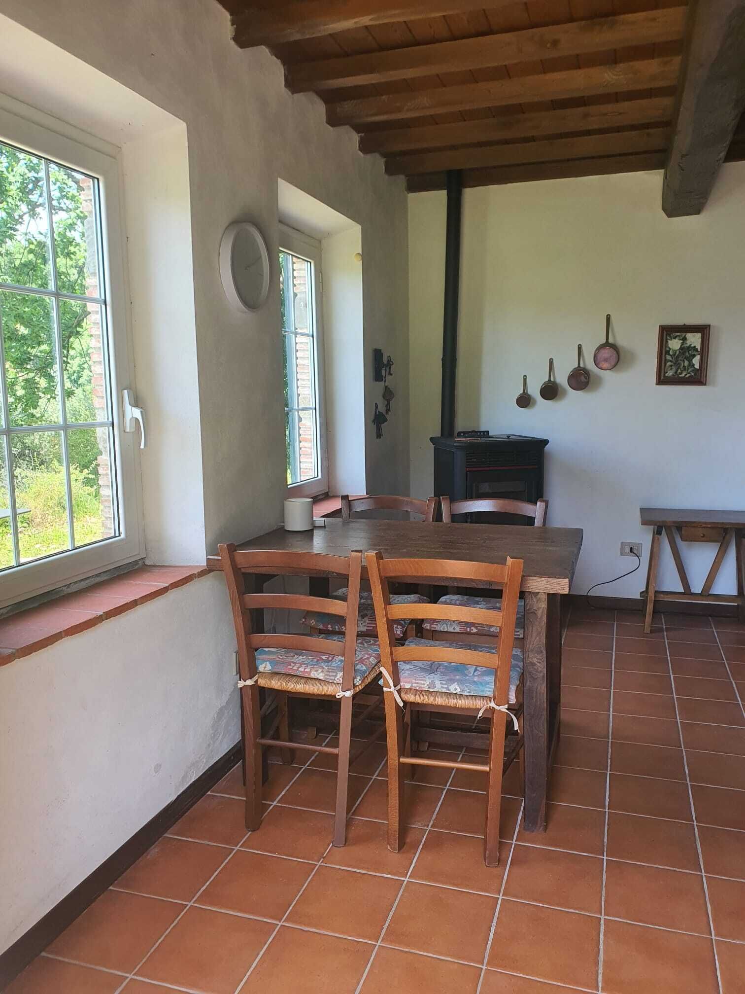 A dining area with a wooden table and chairs, a window with a view of greenery, and a kitchen area with a stove and hanging pots.