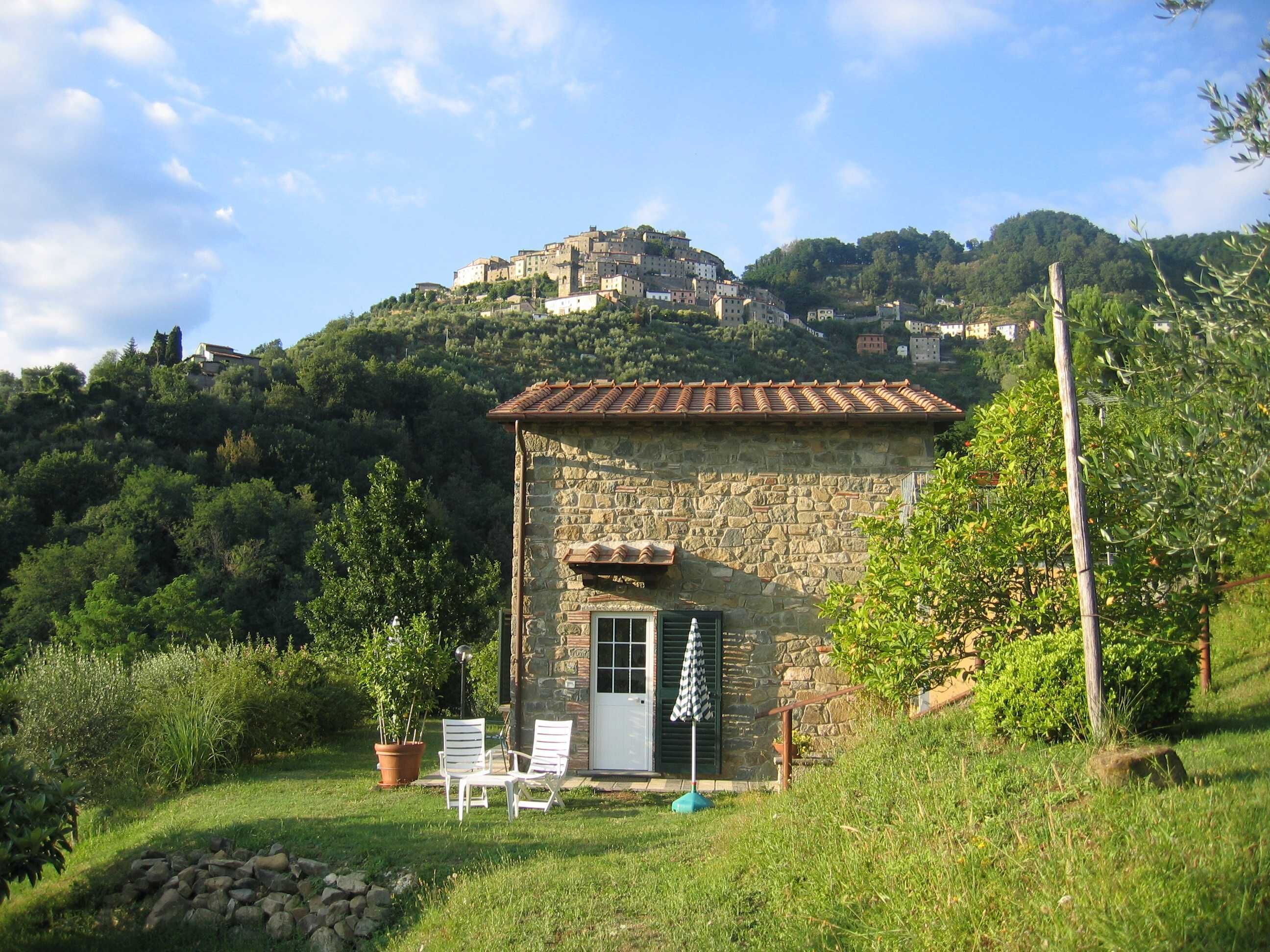 A stone building with a tiled roof, a door, and a small patio with two white chairs and an umbrella.