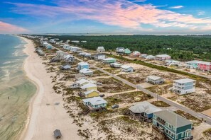 Eine Vogelperspektive eines Küstenwohngebiets mit Häusern entlang des Strandes, einer Straße und bewaldeter Landschaft im Hintergrund.