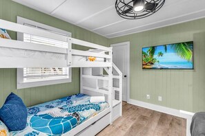 Una cama litera con un edredón azul estampado, un ventilador de techo blanco y un cuadro con una playa en la pared.