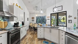 Une cuisine moderne avec des meubles blancs, un grand îlot central et un coin repas offrant une vue sur l’extérieur.