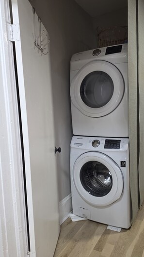 Une machine à laver et un sèche-linge superposés dans une buanderie, avec une porte blanche et un plancher en bois.