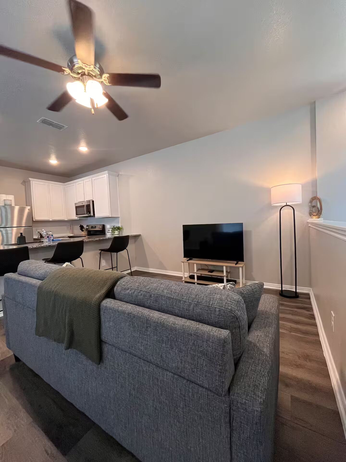 A living room with a gray sofa, a flat-screen TV, a ceiling fan, and a kitchen in the background.