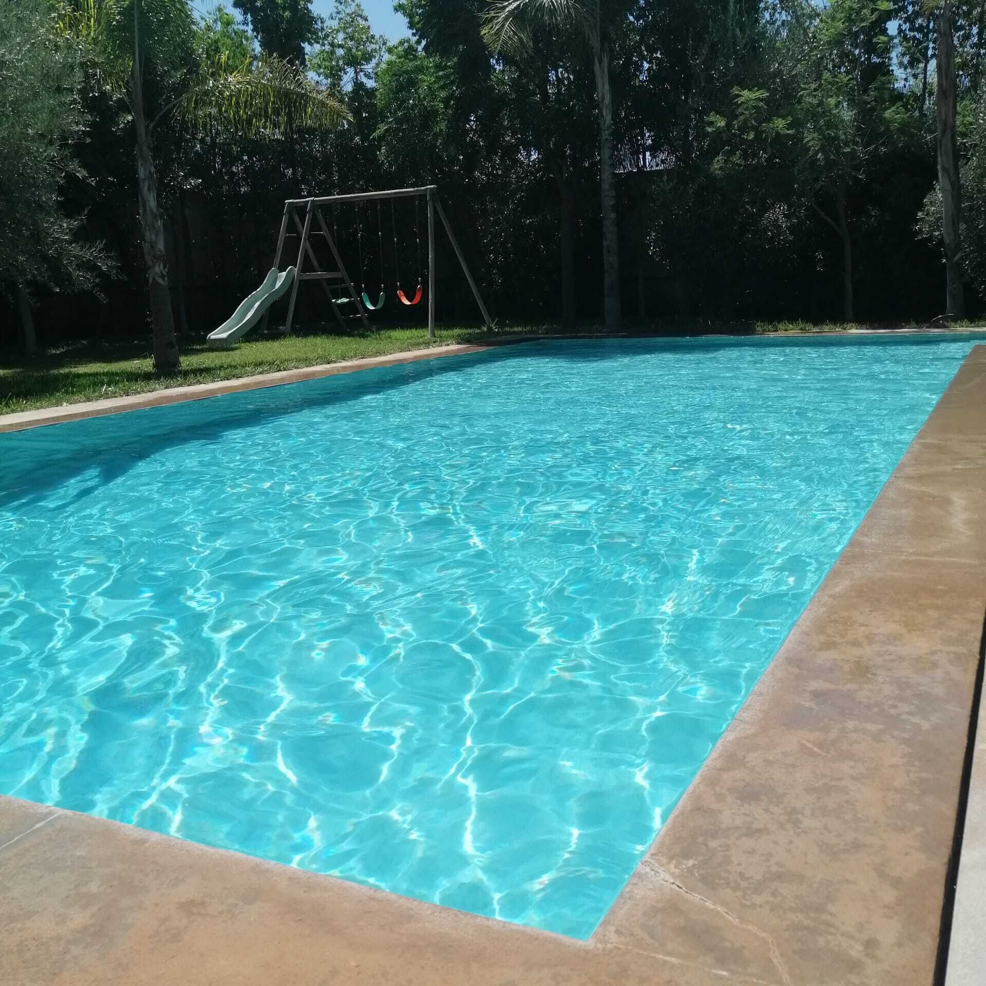 A swimming pool with clear blue water, surrounded by a concrete deck and a grassy area with trees.