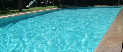 A swimming pool with clear blue water, surrounded by a concrete deck and a grassy area with trees.