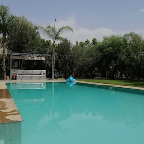 Zwembad met een blauw opblaasbaar speeltje, omgeven door bomen en een overdekt gedeelte.