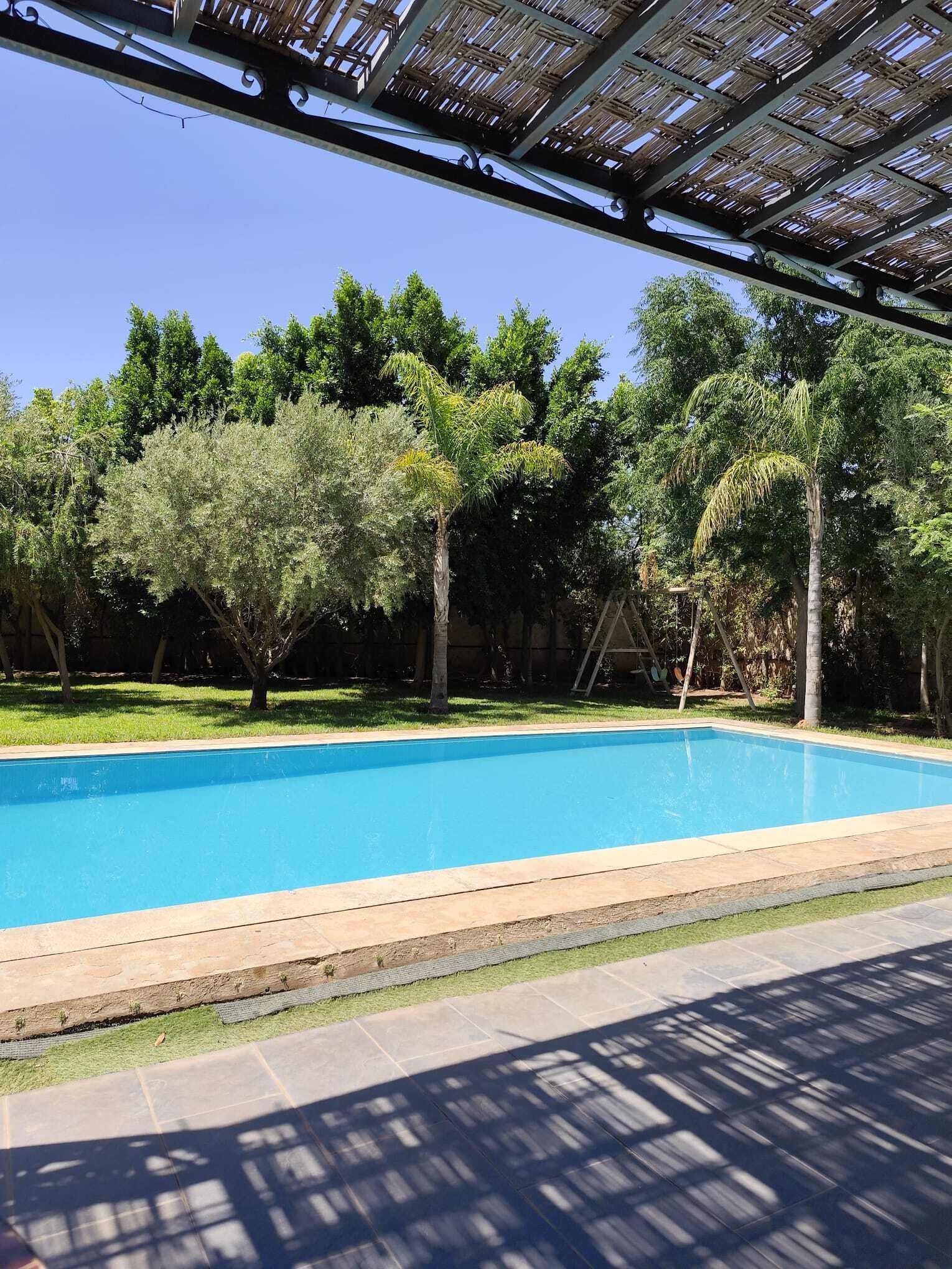 A clear blue swimming pool with a wooden deck edge, surrounded by lush green trees and a shaded area with a thatched roof.