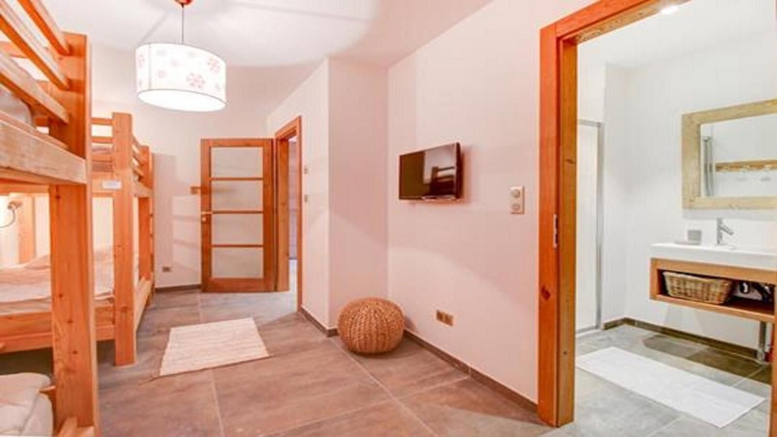 A hotel room with a wooden bunk bed, a small round woven stool, a flat-screen TV mounted on the wall, and a bathroom with a sink and mirror.