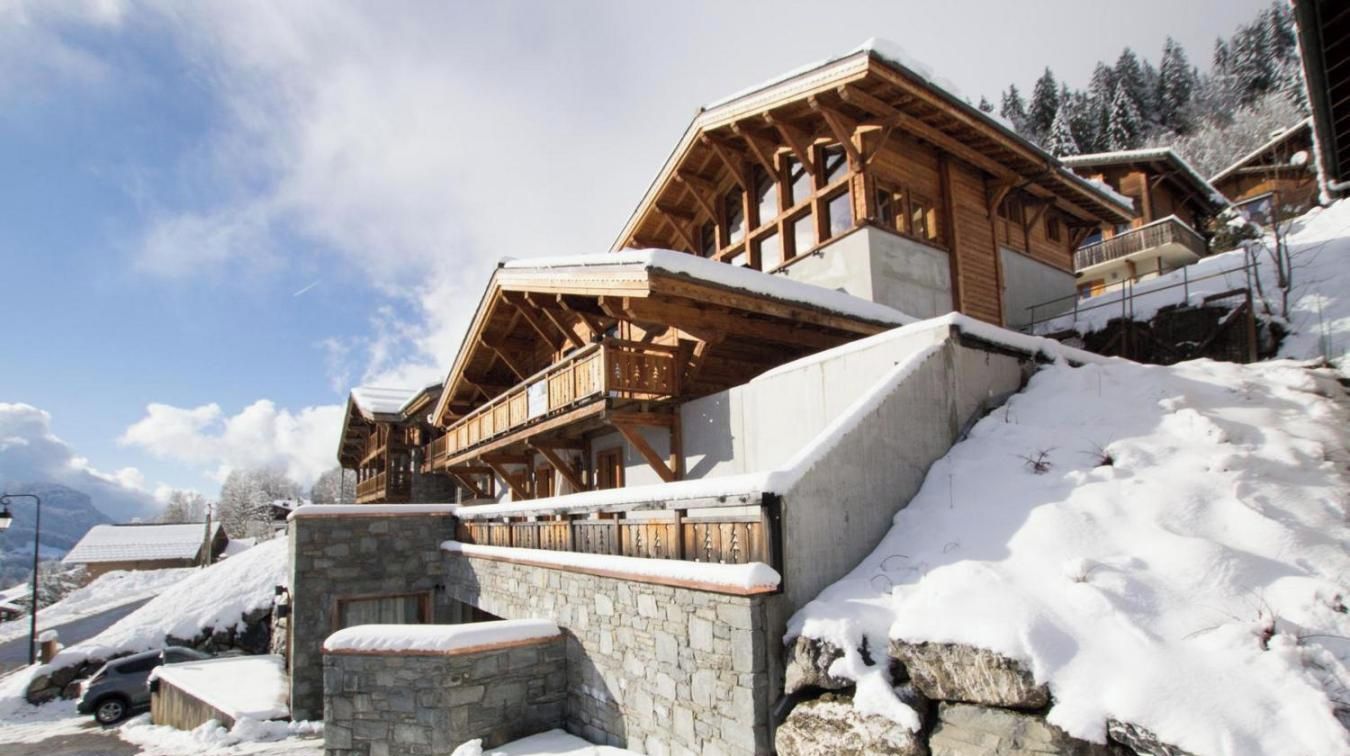 Un chalet en bois, avec des fondations en pierre, se dressant sur un fond de montagnes enneigées.
