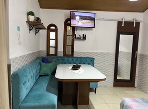 Ein Zimmer mit einem petrolfarbenen Sofa, einem kleinen Esstisch, einem an der Wand befestigten Fernseher, einem Spiegel und einer Holztür.