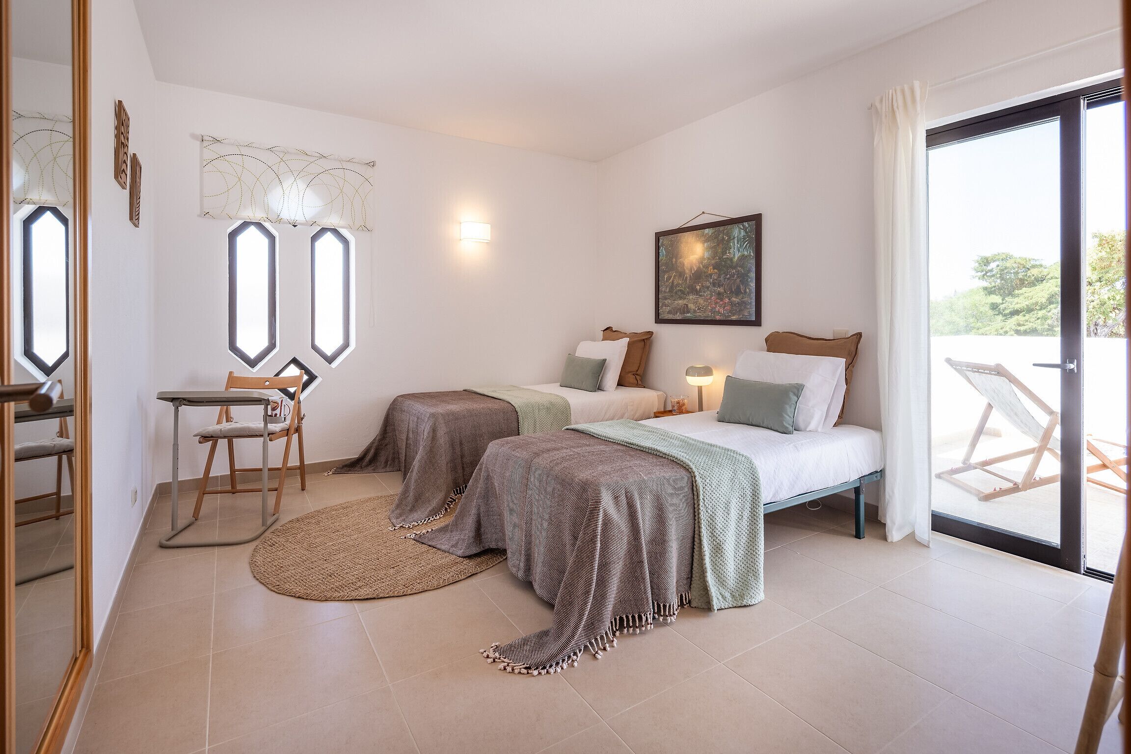 Une chambre avec deux lits, un bureau et une chaise. Il y a une fenêtre avec des rideaux, un tableau au mur et une vue sur l’extérieur.