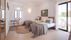 A bedroom with two beds, a desk, and a chair. There is a window with curtains, a painting on the wall, and a view of the outdoors. - CAJI- NEW VILLA NEAR THE BEACH (Albufeira)