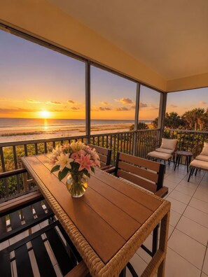 A balcony with a wooden table and chairs, a vase of flowers, and a view of the sunset over the ocean. - Gulffront Paradise at Vacation Villa (Fort Myers Beach)