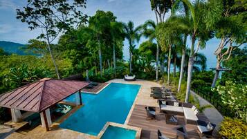 A pool area with lounge chairs and a wooden deck, surrounded by tropical vegetation and overlooking a body of water.