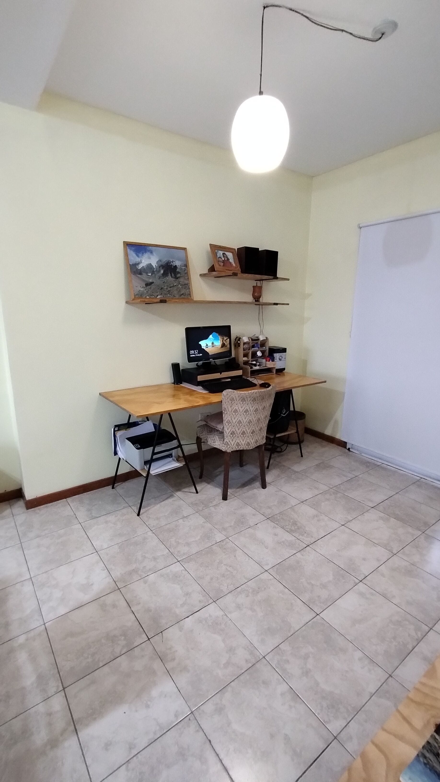 Una oficina en casa con escritorio, computadora, silla, estanterías con libros y un marco de fotos, además de una lámpara colgante.