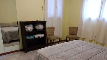 A hotel room with a bed, a wardrobe, two chairs, an air conditioning unit, and a window with curtains.