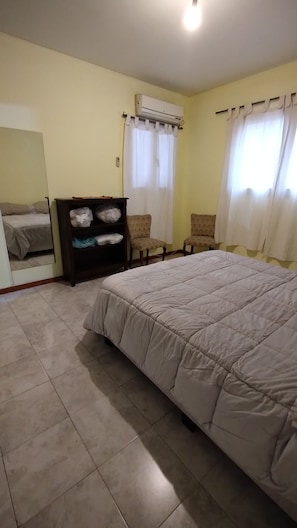 A hotel room with a bed, a wardrobe, two chairs, an air conditioning unit, and a window with curtains.