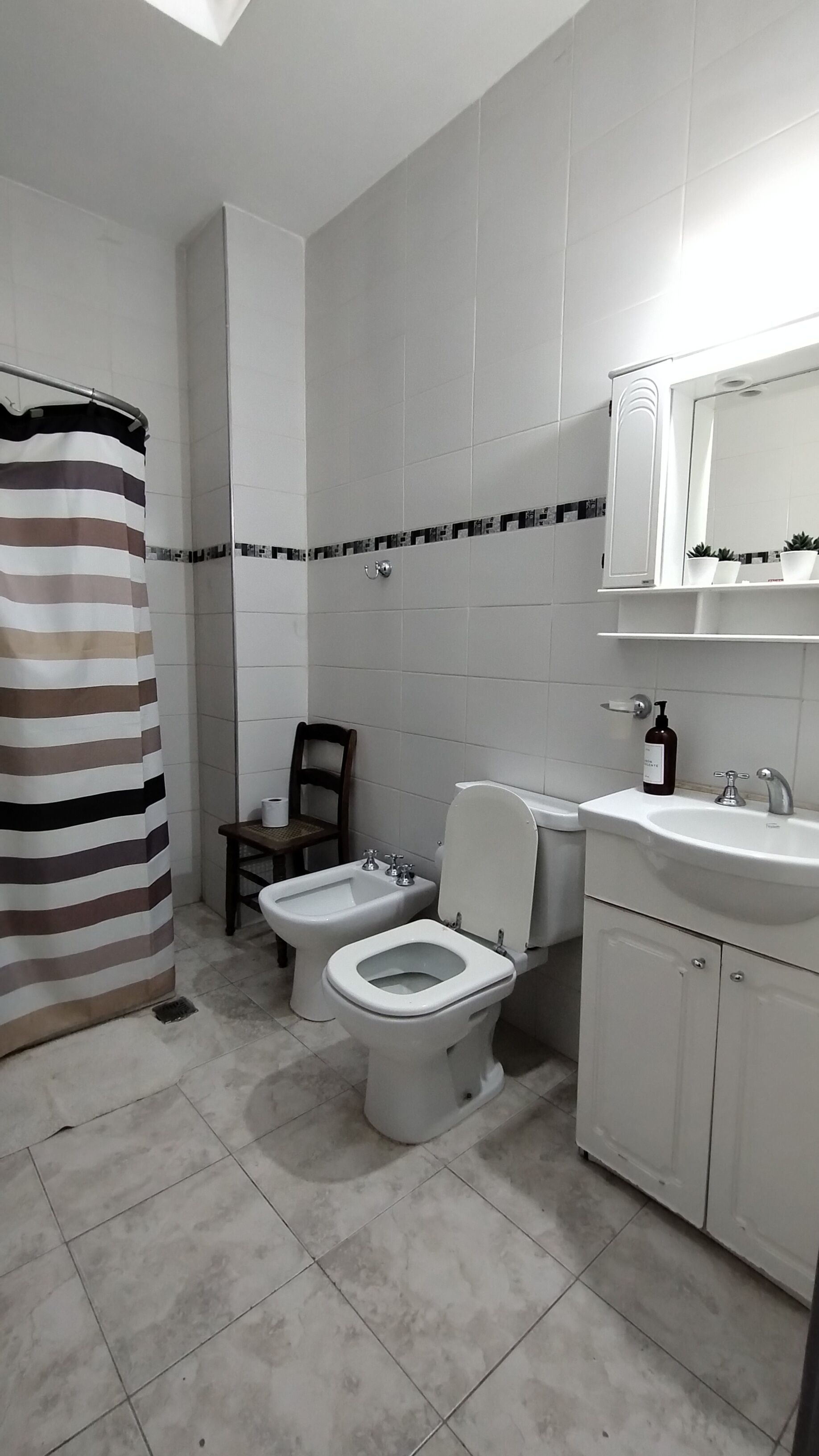 A bathroom with a toilet, bidet, and sink. There is a striped shower curtain and a chair.